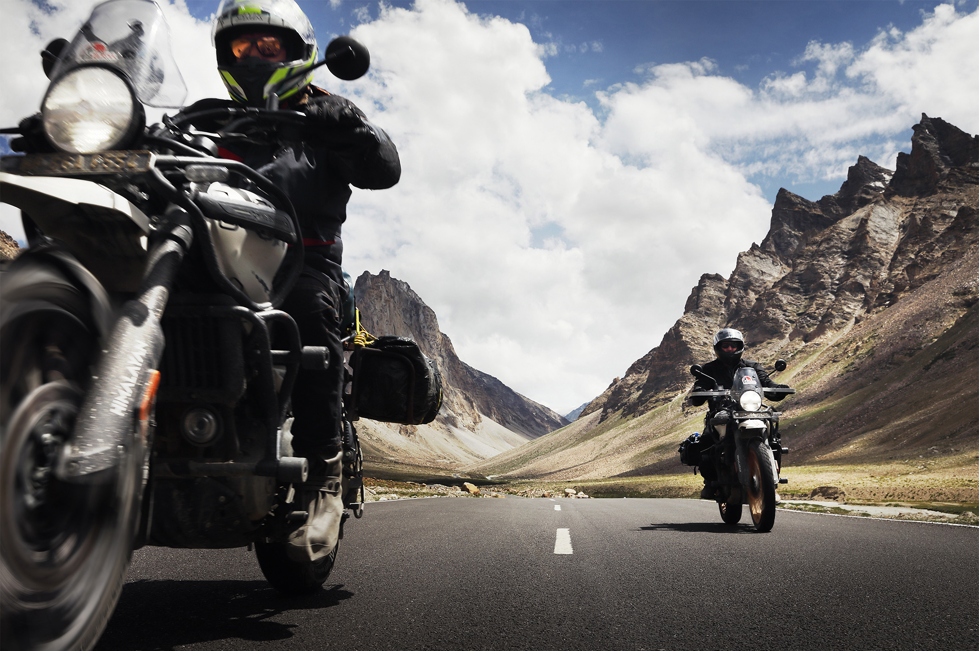 Motorcyclist riding on a mountain road in Ladakh with snow-capped peaks and barren high-altitude desert terrain.
