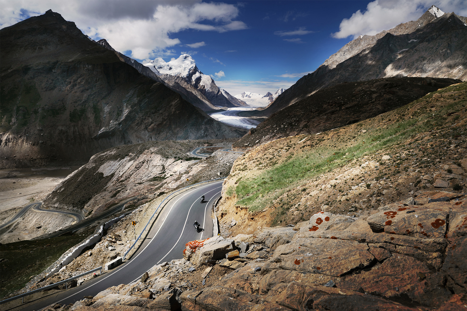 Motorcyclist riding on a mountain road in Ladakh with snow-capped peaks and barren high-altitude desert terrain.
